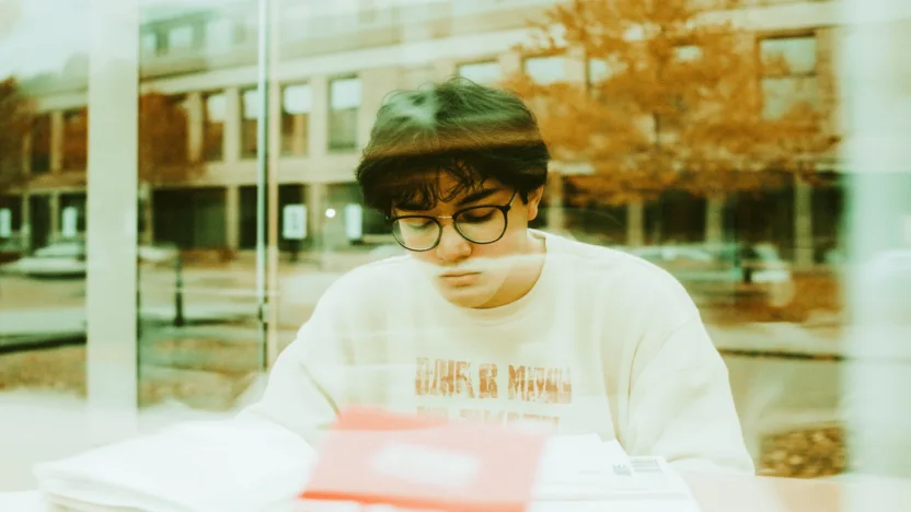 Student rozważający czy rezygnacja ze studiów jest dla niego, przeglądający oficjalne dokumenty na biurku w pokoju akademickim. Widoczna legitymacja studencka i laptop, w tle budynek uniwersytetu za oknem.