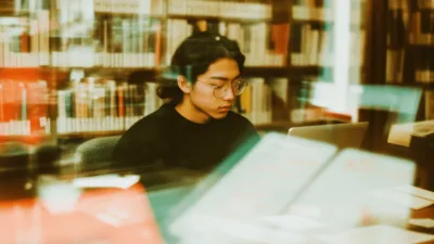 Student w okularach planujący indywidualny tok studiów przy biurku uniwersyteckiej biblioteki, z rozłożonymi książkami, laptopem i kalendarzem z zaznaczonymi terminami.