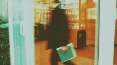 Student w stroju biznesowym wchodzący do nowoczesnego biurowca na praktyki studenckie, trzymający notes i kawę, symbolizujący początek kariery zawodowej.