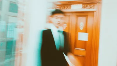 Student stojący przed drzwiami dziekanatu, trzymający w dłoni podanie do dziekana. Formalne otoczenie uczelni z drewnianymi elementami i naturalnym oświetleniem.